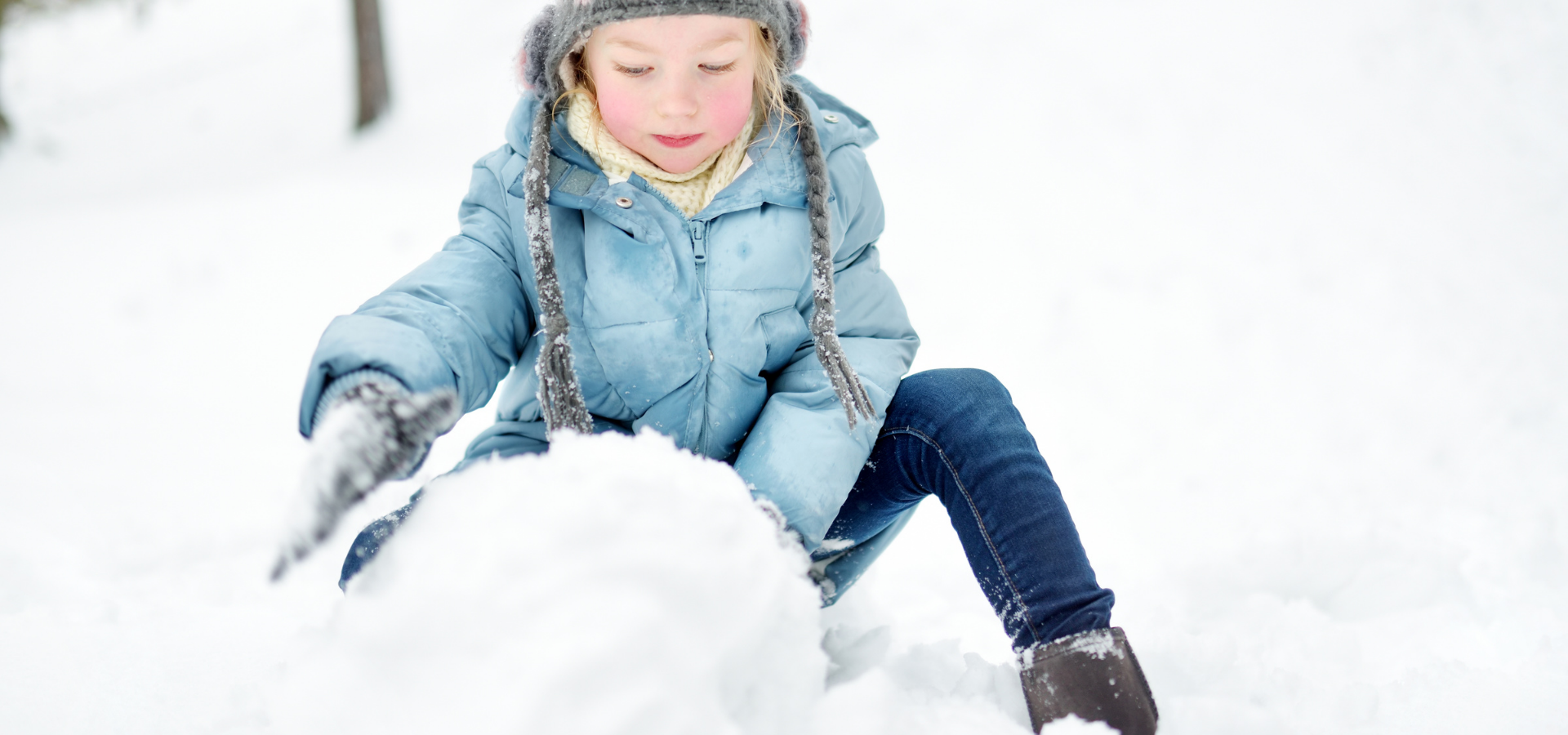 Egy kislány hóban játszik, kék kabátban és sapkában, boldogan épít hóembert.
